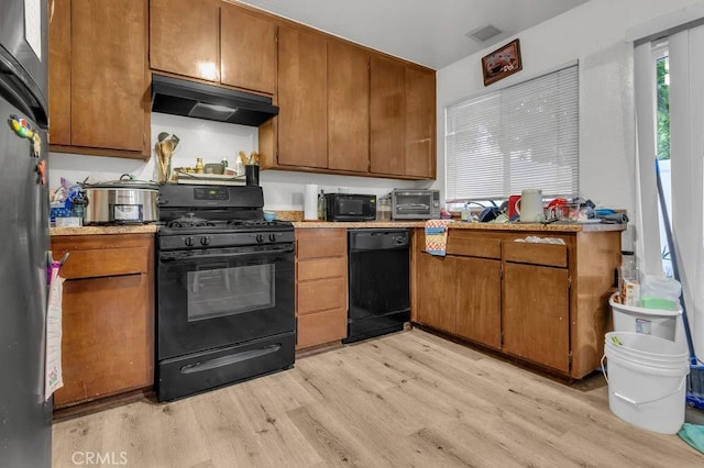 kitchen with a toaster, brown cabinetry, under cabinet range hood, black appliances, and light wood finished floors