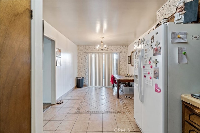 kitchen featuring light tile patterned floors, a notable chandelier, white refrigerator with ice dispenser, baseboards, and wallpapered walls