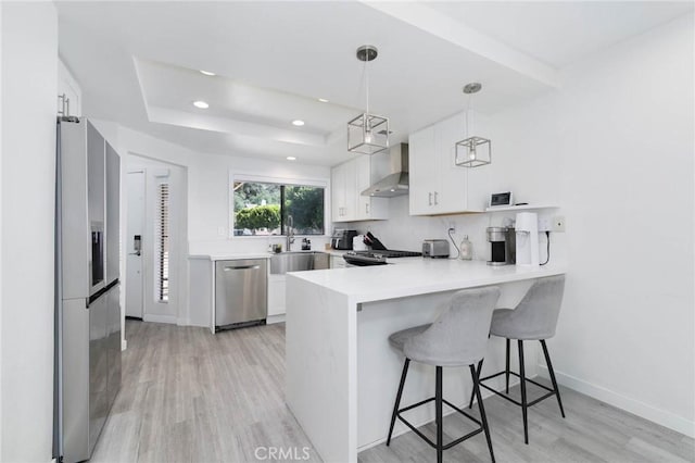 kitchen featuring appliances with stainless steel finishes, wall chimney exhaust hood, a peninsula, light wood-type flooring, and a tray ceiling