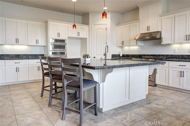 kitchen with stove, under cabinet range hood, white cabinets, pendant lighting, and a center island with sink