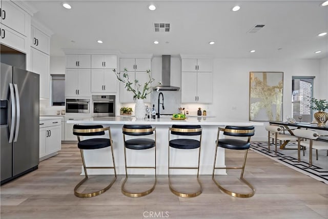 kitchen with wall chimney range hood, a kitchen island with sink, visible vents, light wood-type flooring, and stainless steel appliances