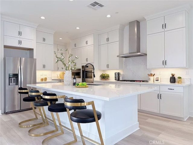 kitchen featuring stainless steel refrigerator with ice dispenser, visible vents, light wood finished floors, wall chimney exhaust hood, and an island with sink