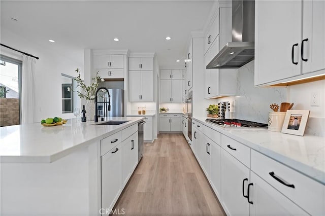 kitchen with wall chimney range hood, white cabinets, appliances with stainless steel finishes, a sink, and light wood finished floors