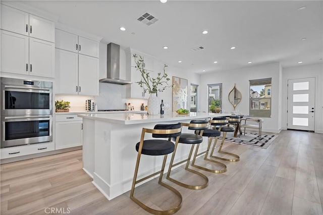 kitchen featuring stainless steel double oven, visible vents, a center island with sink, light wood-style floors, and wall chimney exhaust hood