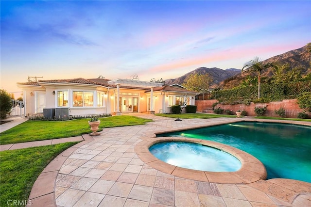 pool at dusk with a fenced backyard, a mountain view, a yard, french doors, and a pool with connected hot tub