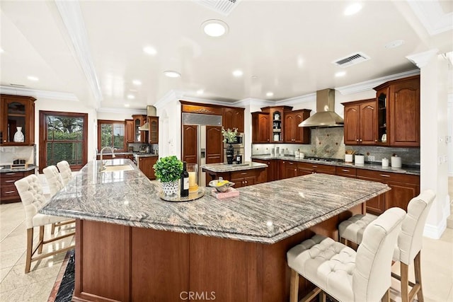 kitchen with wall chimney range hood, visible vents, paneled built in fridge, a kitchen bar, and a large island