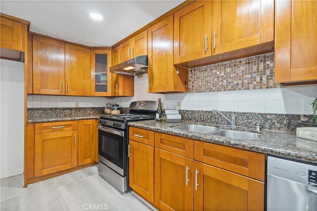 kitchen with a sink, brown cabinets, stainless steel appliances, under cabinet range hood, and dark stone countertops