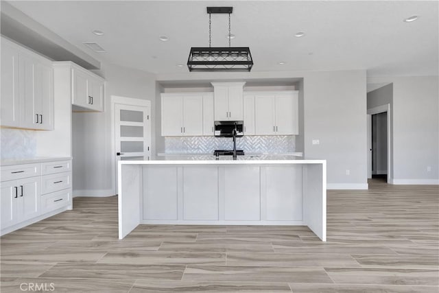 kitchen with a center island with sink, light countertops, stainless steel microwave, white cabinets, and backsplash