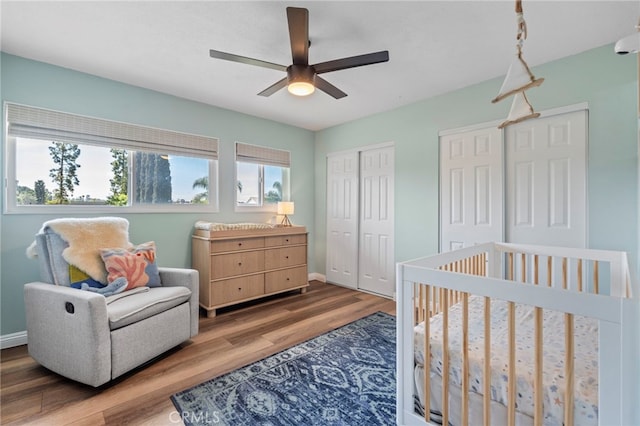 bedroom featuring ceiling fan, a nursery area, baseboards, wood finished floors, and multiple closets