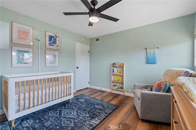 bedroom featuring wood finished floors, a nursery area, baseboards, visible vents, and a ceiling fan