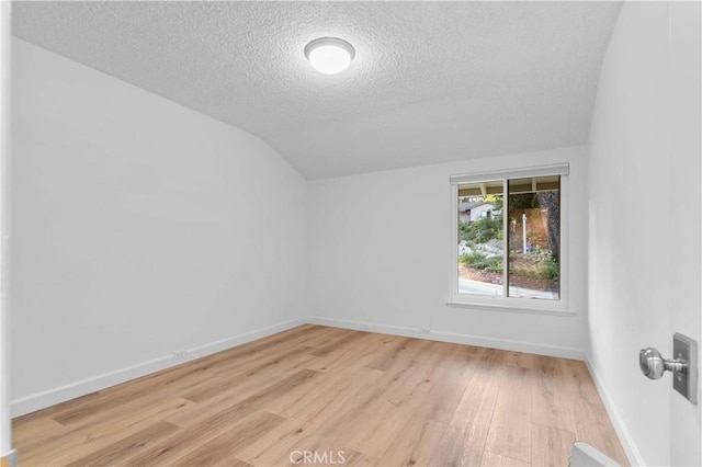 empty room with a textured ceiling, light wood-style flooring, vaulted ceiling, and baseboards