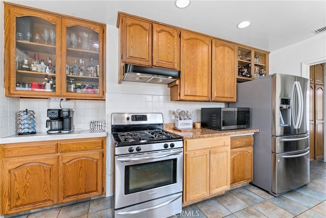 kitchen with tasteful backsplash, glass insert cabinets, stainless steel appliances, under cabinet range hood, and light countertops