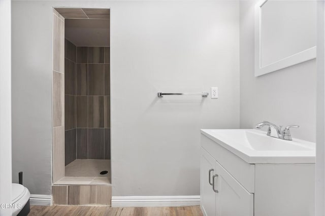 bathroom with a tile shower, baseboards, toilet, wood finished floors, and vanity