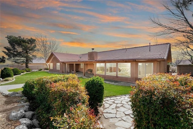 back of house at dusk featuring a patio, a lawn, a tiled roof, a chimney, and fence