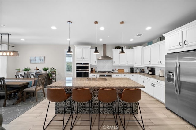 kitchen with wall chimney range hood, tasteful backsplash, stainless steel appliances, and a center island with sink