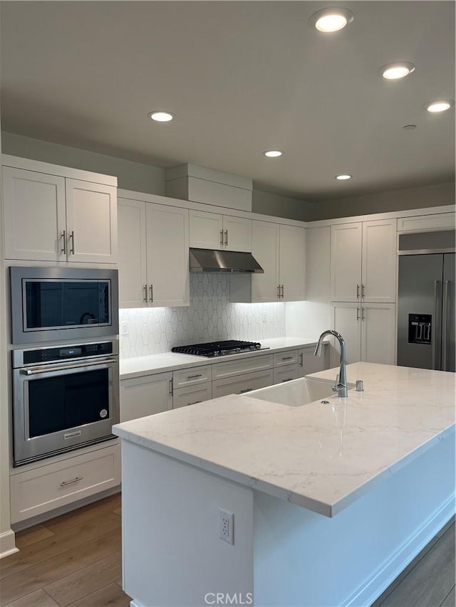 kitchen with decorative backsplash, a sink, built in appliances, under cabinet range hood, and wood finished floors