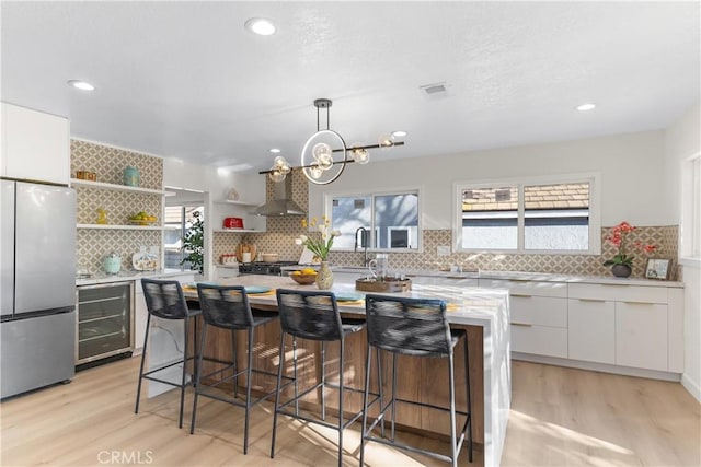 kitchen featuring freestanding refrigerator, open shelves, beverage cooler, wall chimney exhaust hood, and white cabinetry