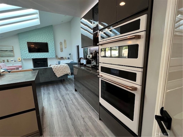 kitchen featuring lofted ceiling, double oven, modern cabinets, a large fireplace, and light wood-style flooring
