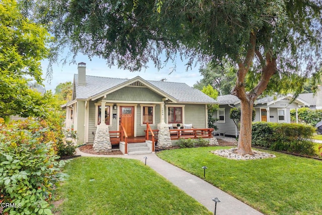 bungalow-style house featuring a shingled roof, covered porch, a chimney, and a front yard