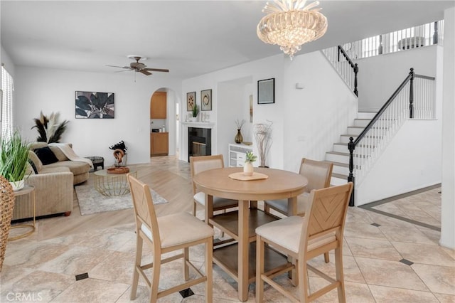 dining area featuring arched walkways, a glass covered fireplace, ceiling fan with notable chandelier, stairs, and light tile patterned floors