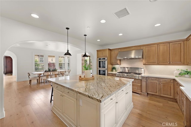kitchen featuring stainless steel double oven, light stone counters, ventilation hood, arched walkways, and visible vents