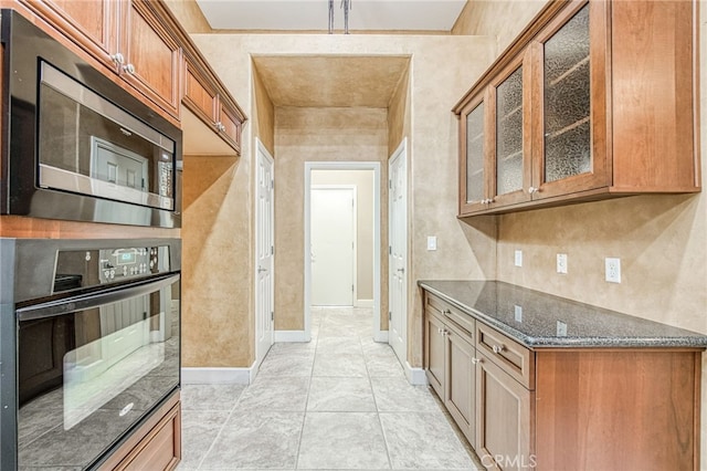 kitchen with dark stone counters, stainless steel microwave, glass insert cabinets, black oven, and brown cabinetry