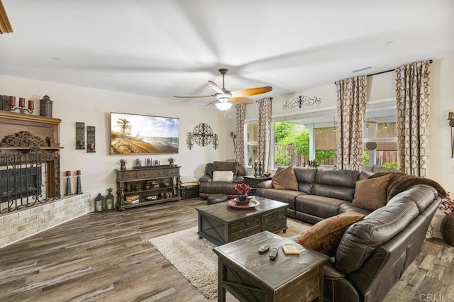 living area with a brick fireplace, a ceiling fan, visible vents, and wood finished floors