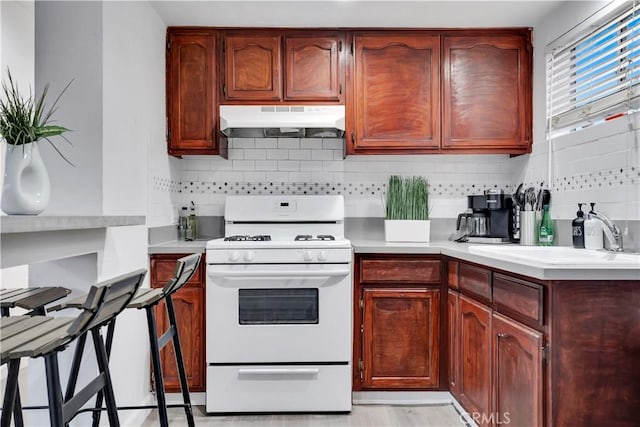 kitchen with white gas range oven, under cabinet range hood, light countertops, backsplash, and a sink