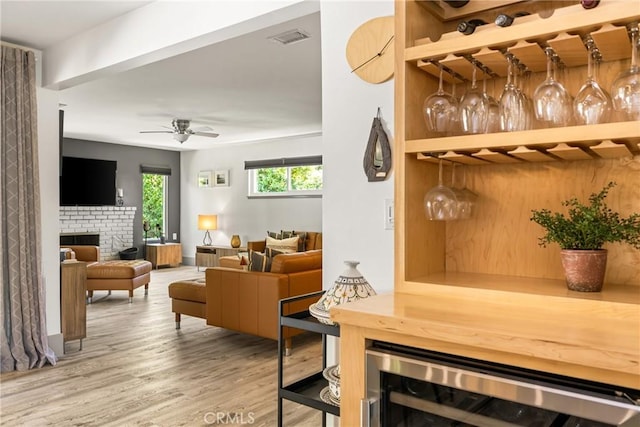 interior space with beverage cooler, ceiling fan, visible vents, a brick fireplace, and wood finished floors