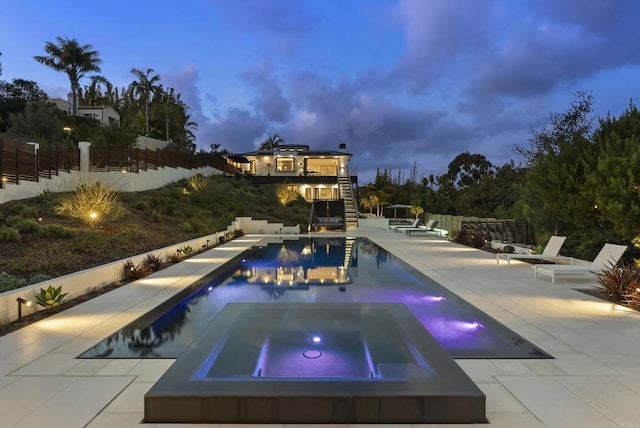 pool at dusk with fence, an outdoor pool, a patio, and an in ground hot tub
