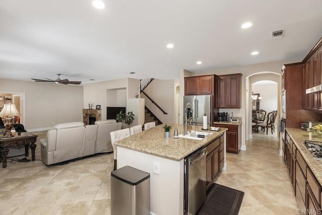 kitchen with a kitchen island with sink, a sink, ceiling fan, arched walkways, and appliances with stainless steel finishes