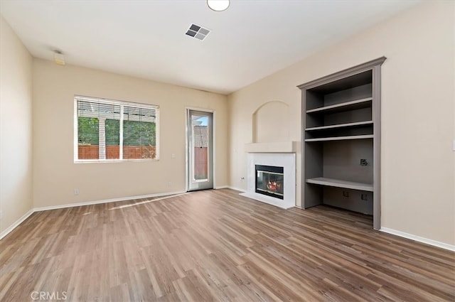 unfurnished living room featuring a fireplace with flush hearth, built in shelves, visible vents, baseboards, and wood finished floors