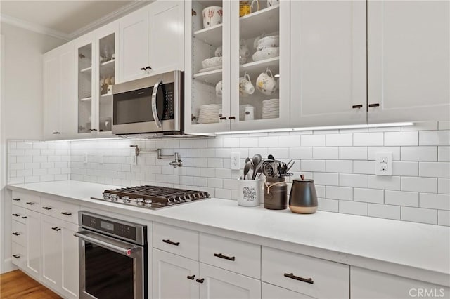 kitchen with crown molding, light countertops, stainless steel appliances, white cabinetry, and glass insert cabinets