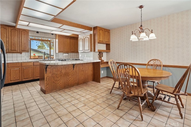 kitchen with stainless steel gas stovetop, brown cabinetry, a peninsula, an inviting chandelier, and tile counters