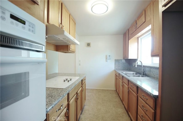 kitchen with light stone countertops, a sink, baseboards, under cabinet range hood, and white appliances