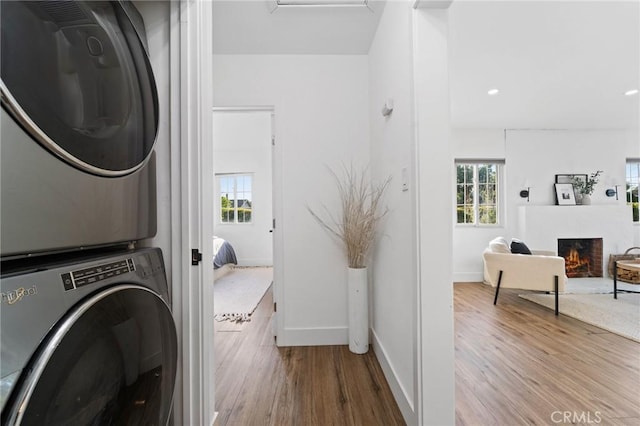 laundry area featuring laundry area, wood finished floors, baseboards, stacked washing maching and dryer, and a warm lit fireplace