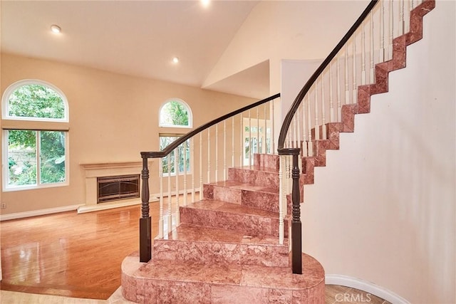 stairway featuring wood finished floors, baseboards, high vaulted ceiling, a glass covered fireplace, and recessed lighting