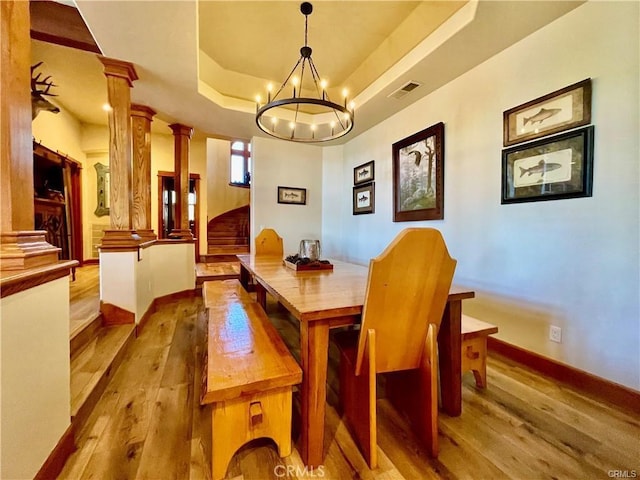 dining room featuring visible vents, wood finished floors, a raised ceiling, a notable chandelier, and ornate columns