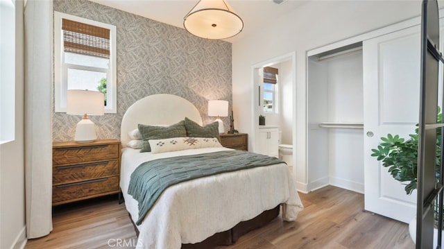 bedroom featuring an accent wall, baseboards, wallpapered walls, ensuite bathroom, and wood finished floors