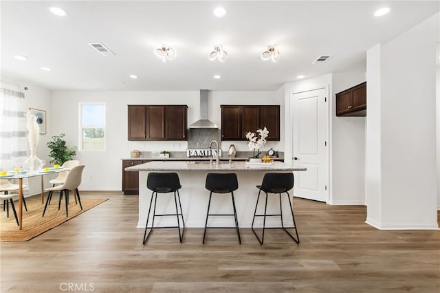 kitchen featuring wall chimney range hood, dark brown cabinets, visible vents, and a kitchen island with sink