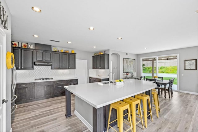 kitchen featuring appliances with stainless steel finishes, a sink, backsplash, under cabinet range hood, and a center island with sink