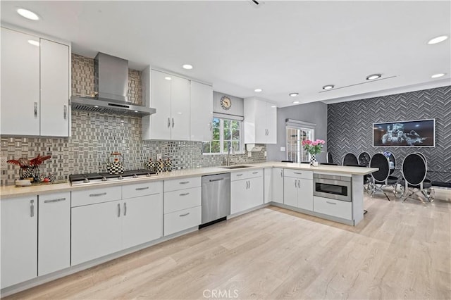 kitchen featuring wall chimney range hood, appliances with stainless steel finishes, light countertops, a peninsula, and light wood-type flooring