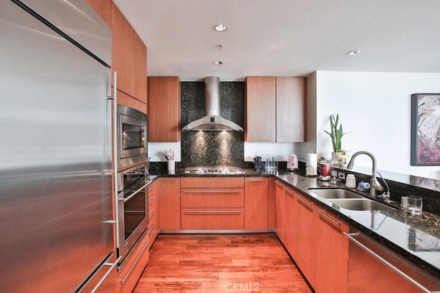 kitchen with built in appliances, dark stone counters, wall chimney exhaust hood, a sink, and light wood-style flooring
