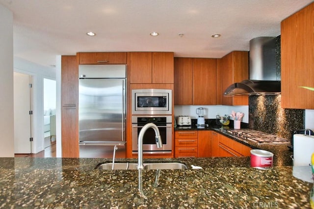 kitchen featuring dark stone counters, wall chimney exhaust hood, built in appliances, and brown cabinets