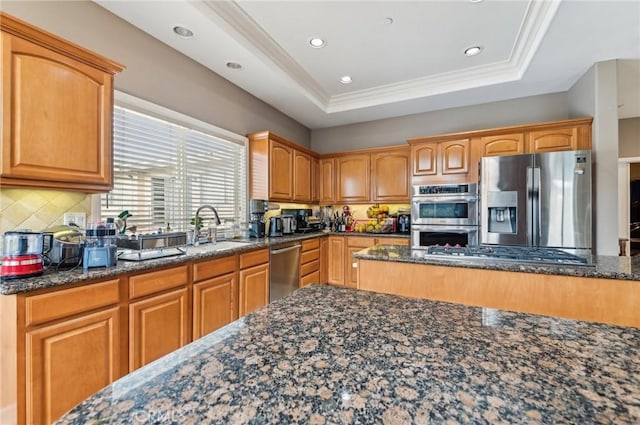 kitchen with a sink, dark stone counters, stainless steel appliances, tasteful backsplash, and a tray ceiling