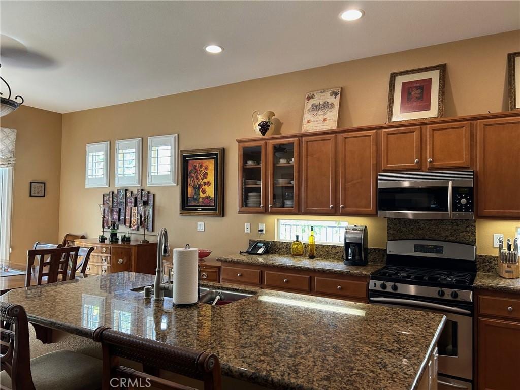 kitchen featuring glass insert cabinets, a kitchen bar, dark stone counters, brown cabinets, and stainless steel appliances