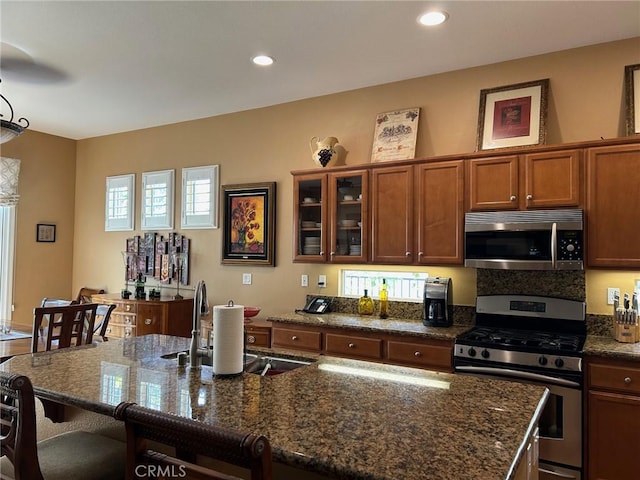 kitchen featuring glass insert cabinets, a kitchen bar, dark stone counters, brown cabinets, and stainless steel appliances