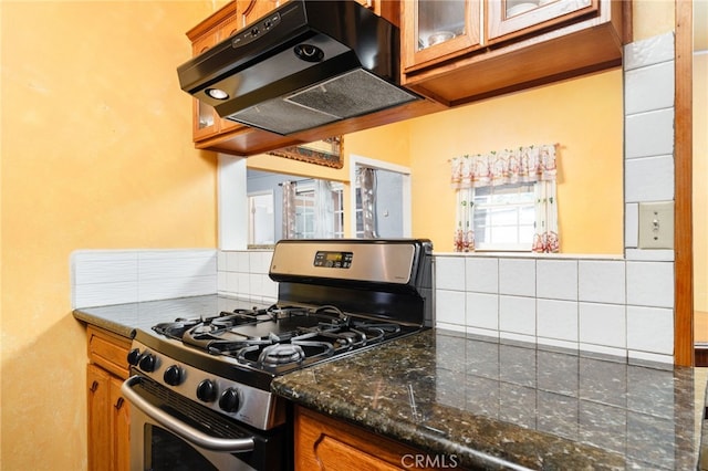kitchen featuring glass insert cabinets, under cabinet range hood, brown cabinets, stainless steel range with gas stovetop, and backsplash