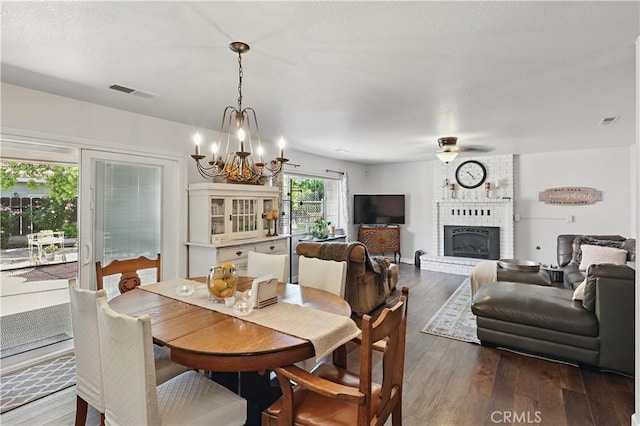 dining area with a brick fireplace, ceiling fan with notable chandelier, visible vents, and dark wood finished floors