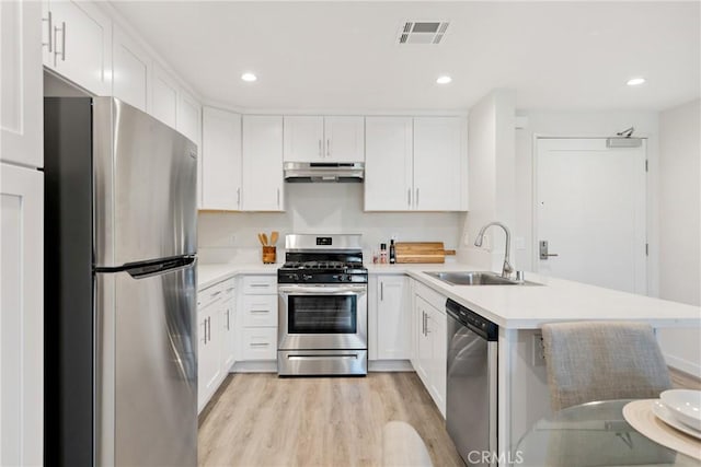 kitchen with a peninsula, under cabinet range hood, a sink, light wood finished floors, and stainless steel appliances